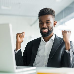 Happy african-american businessman in suit looking at laptop excited by good news online, black man winner sitting at office desk achieved goal raising hands celebrating business success win result